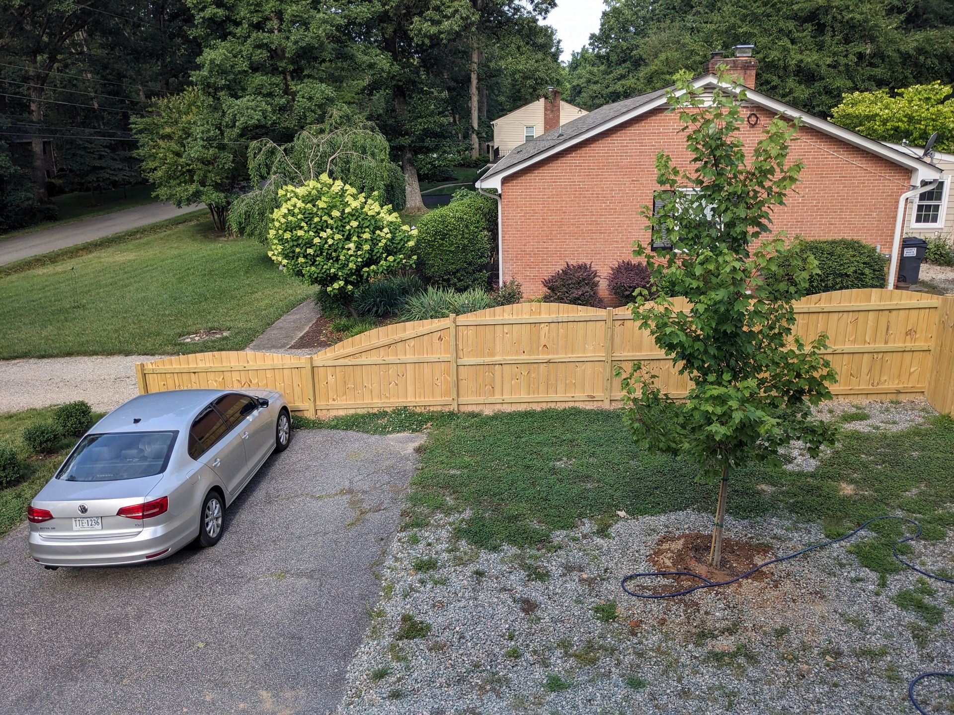 Silver car parked on gravel driveway, next to a wooden fence and brick house. Green trees in the background.