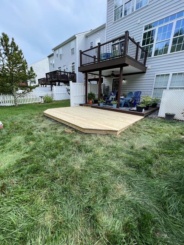 A wooden deck in backyard with a multi-story home. Green grass and a white fence surround the deck.