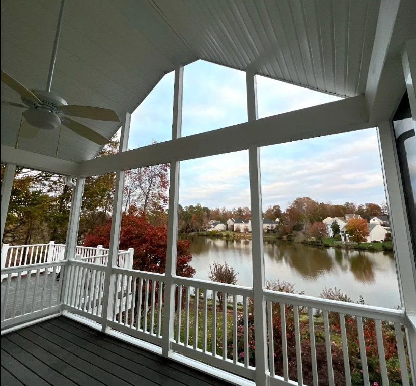 Screened porch with lake view: white railings, dark floor, trees with fall foliage, and a ceiling fan.