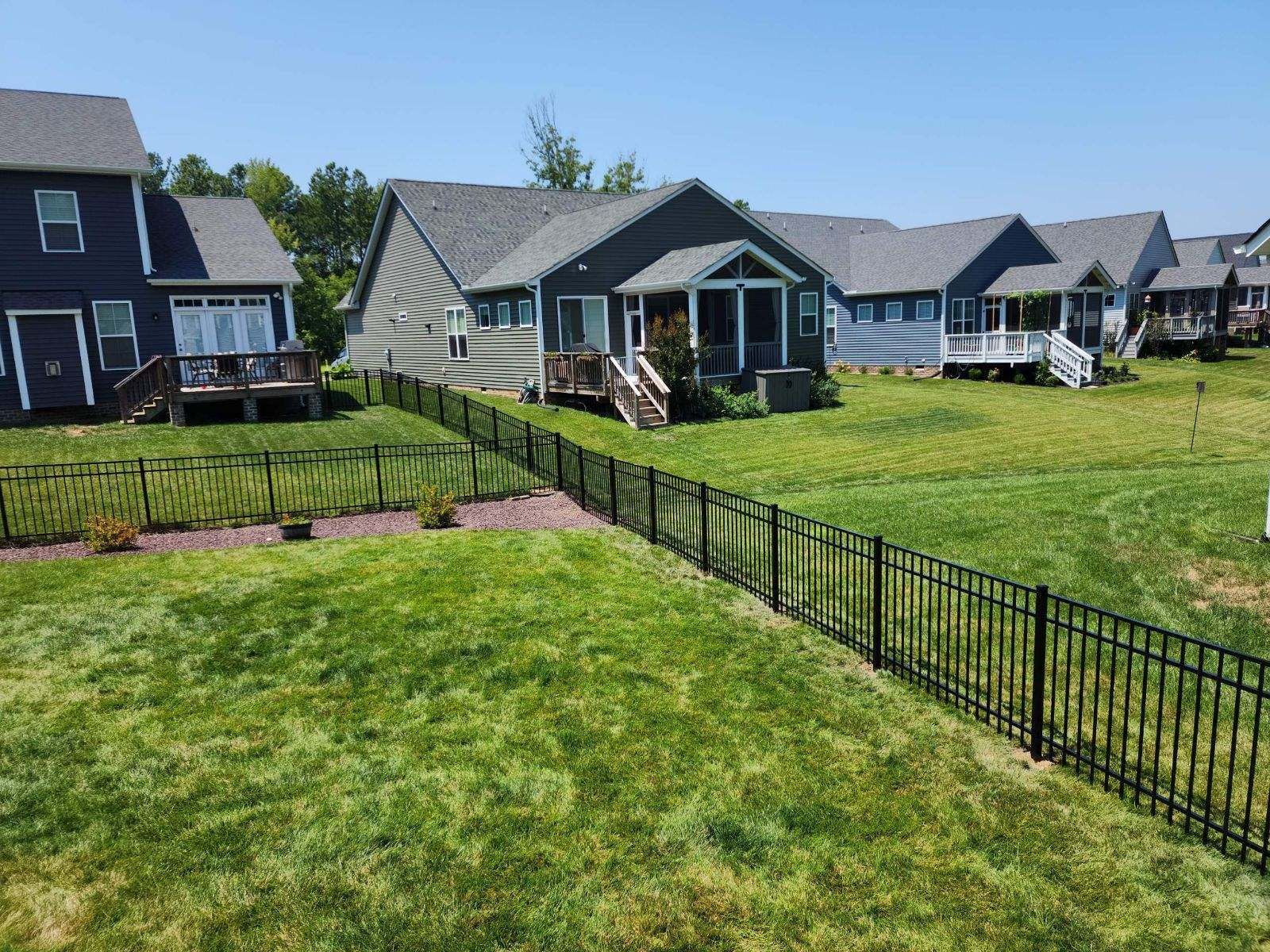 Black fence bordering backyards with houses, green grass, and a blue sky.