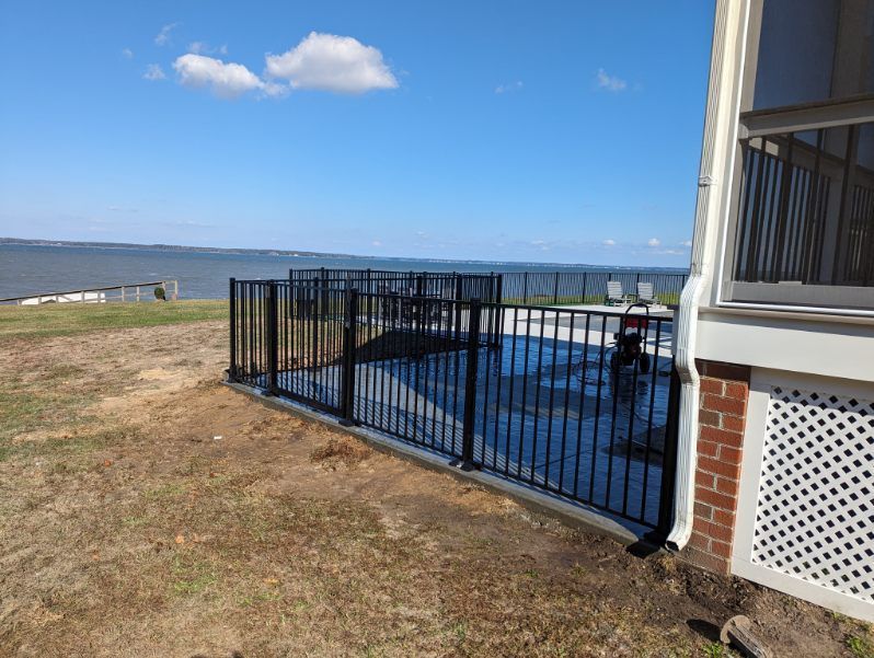 Black fenced patio overlooking water, adjacent to a house with a blue sky.