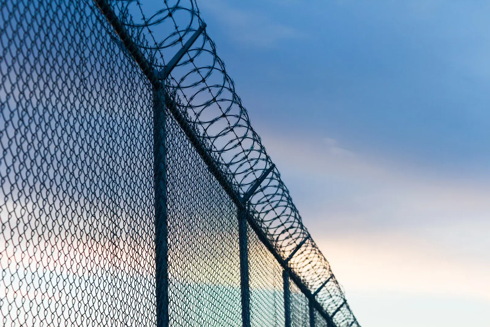 Chain-link fence topped with barbed wire against a blue and pink sky.