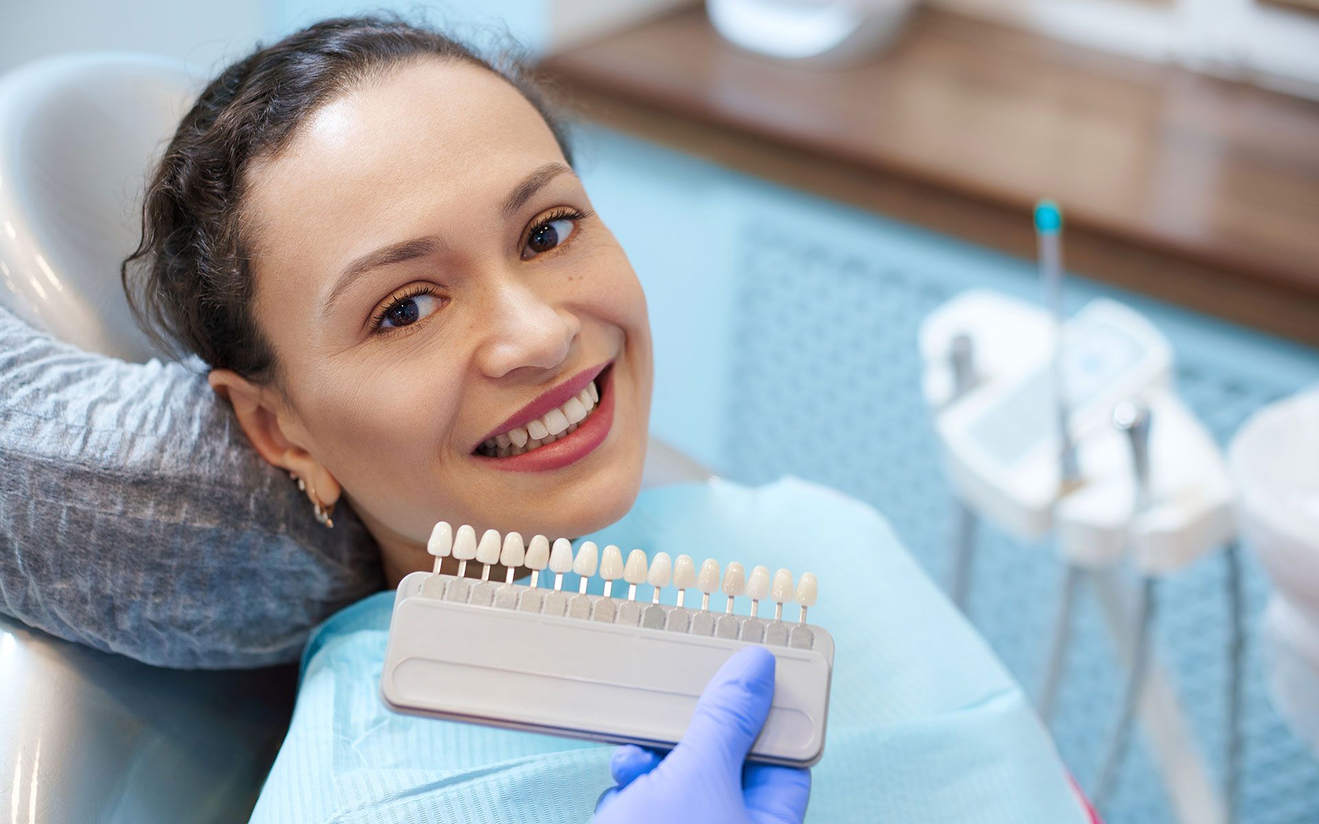 A woman is sitting in a dental chair while a dentist holds a tooth color chart.