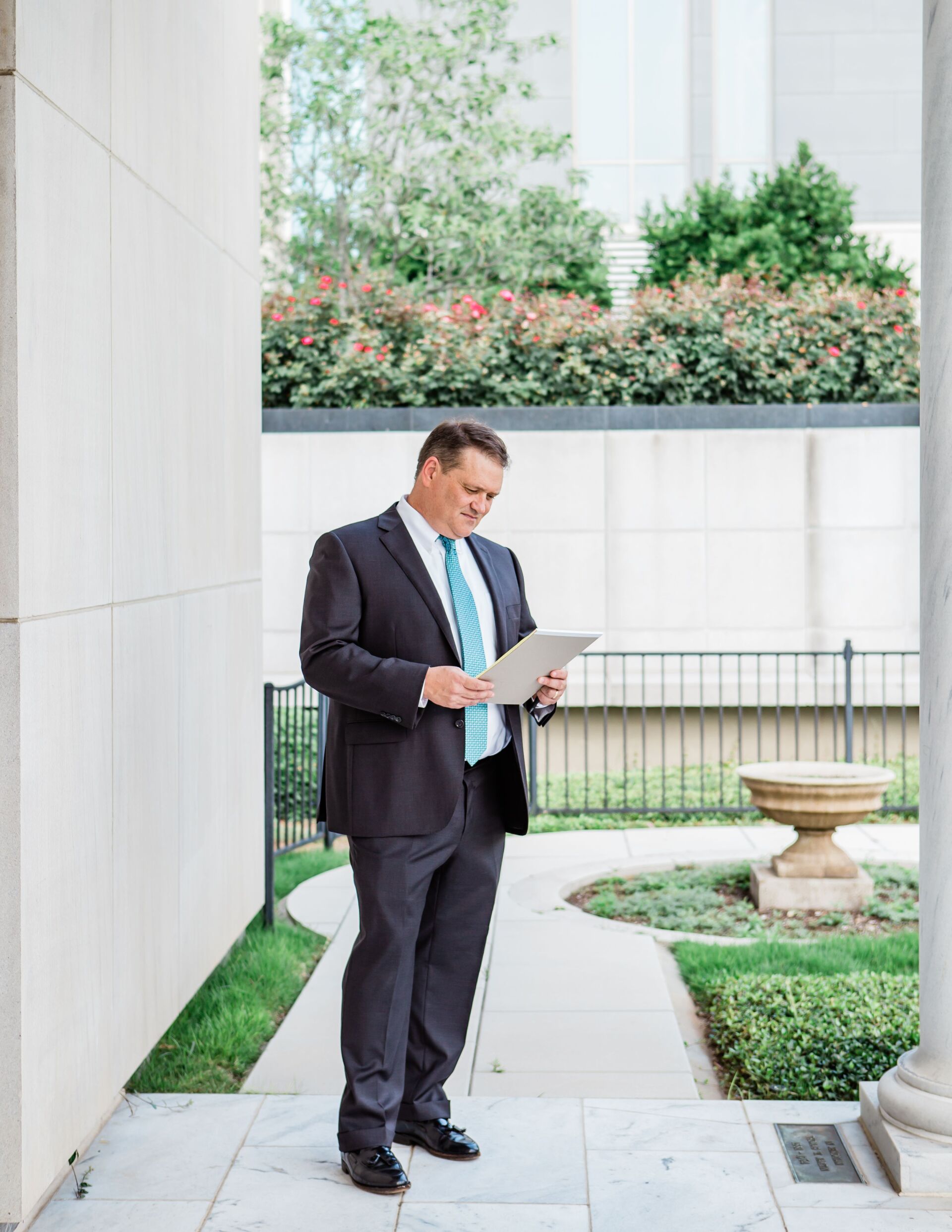 A man in a suit and tie is standing on a sidewalk looking at a piece of paper.