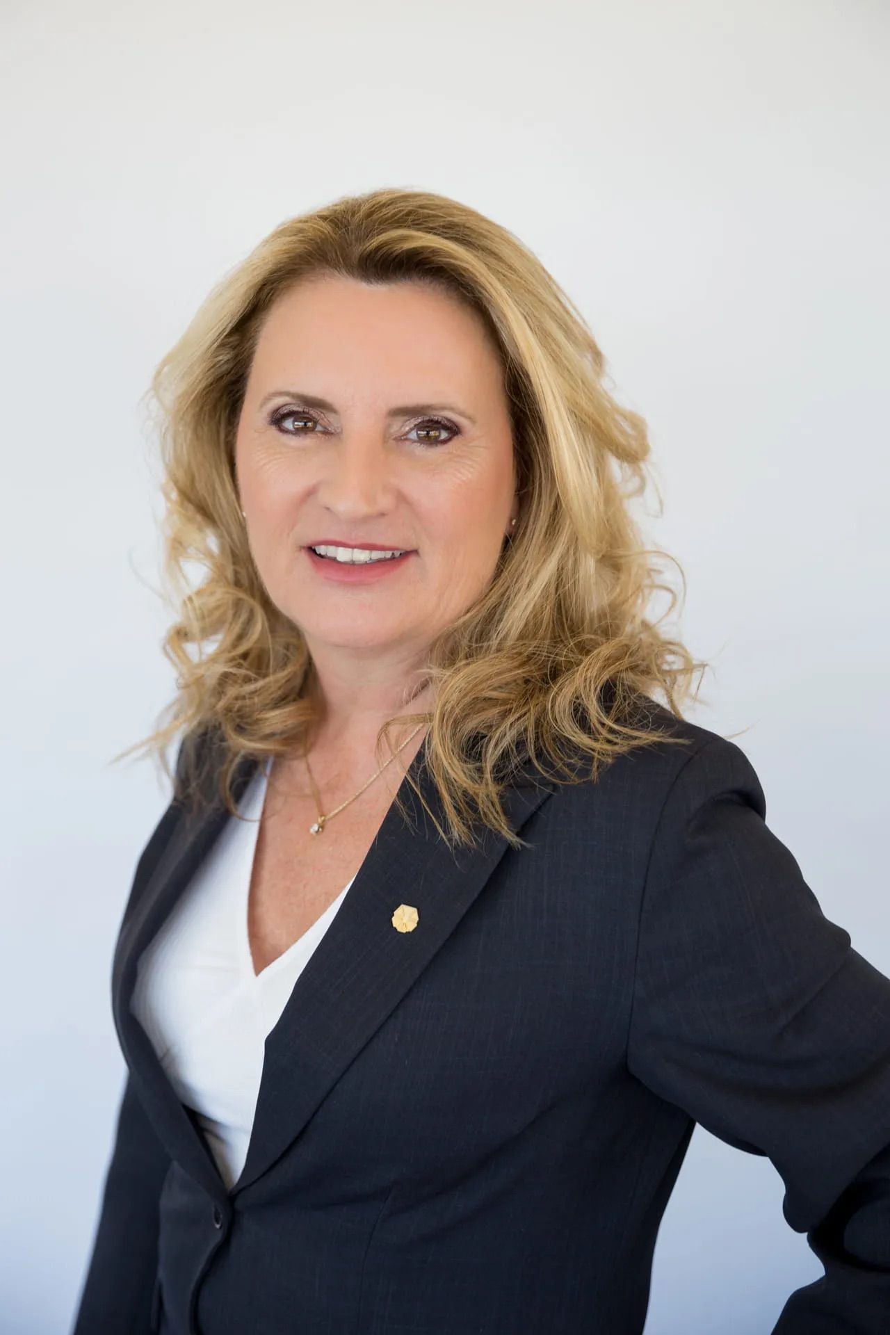 Femme en blazer foncé et chemise blanche, souriante, collier et broche en or, sur fond blanc.