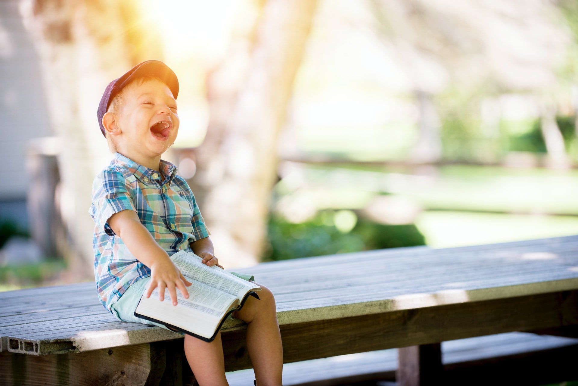 A little boy is sitting on a picnic table holding a bible and laughing.