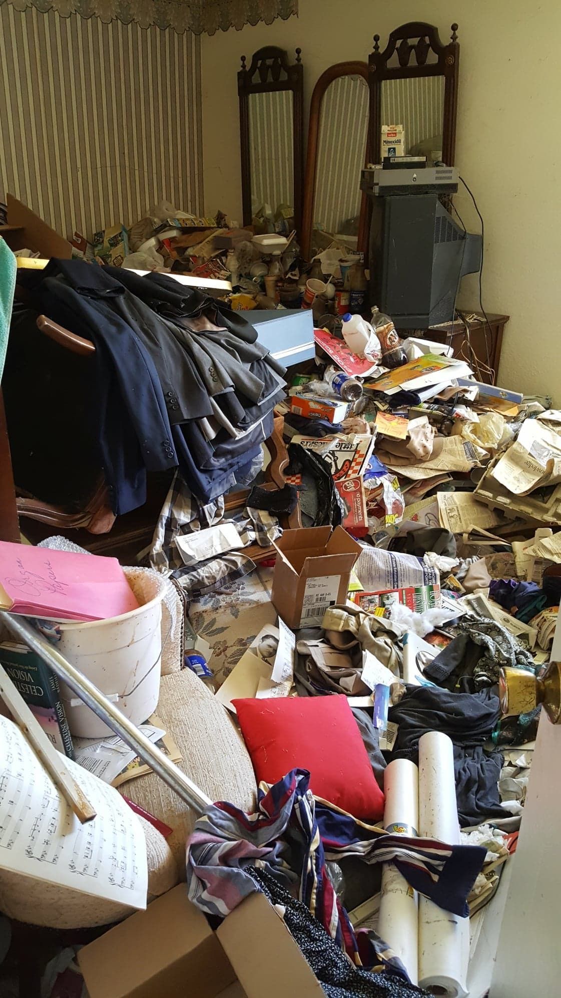 Cluttered room filled with stacks of papers, clothes, and household items, with mirrors and furniture along the wall.