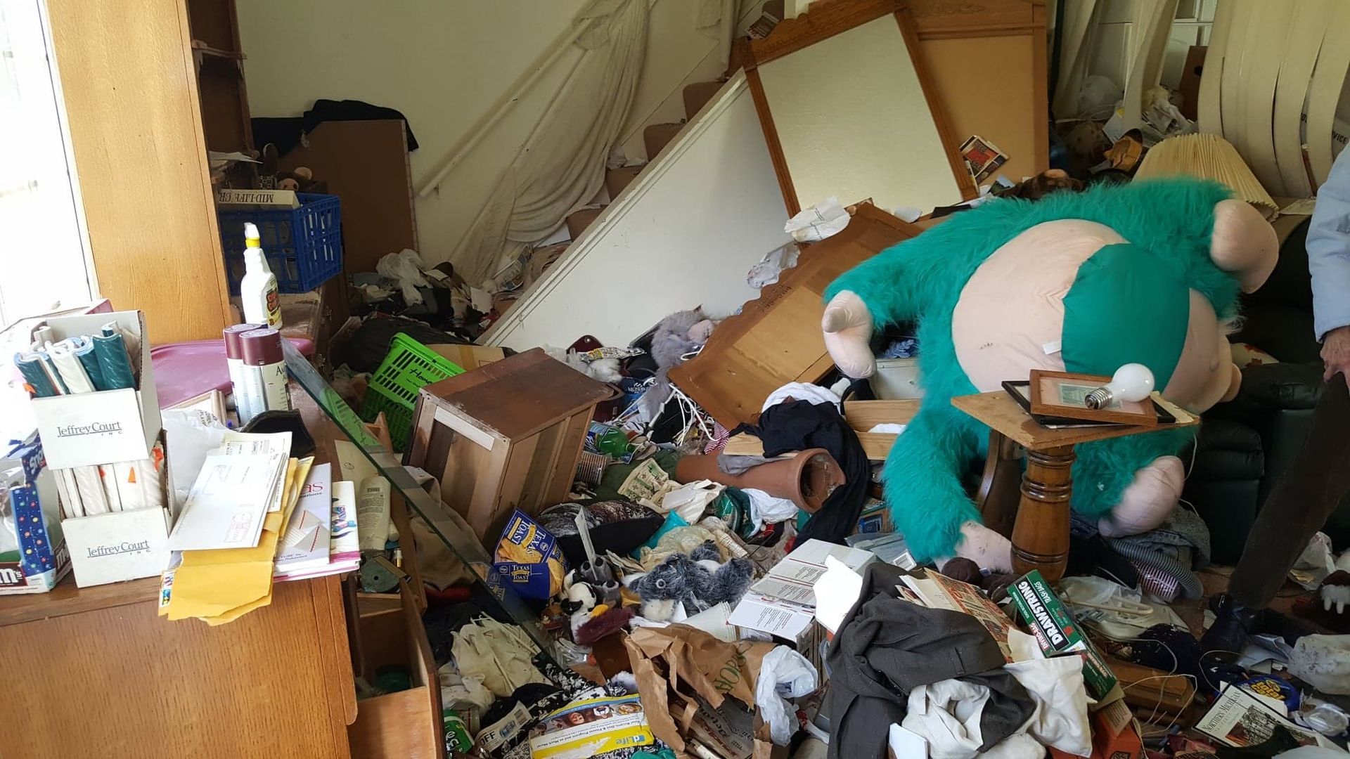 Person in green bends over amid a heavily cluttered room filled with scattered clothes and debris