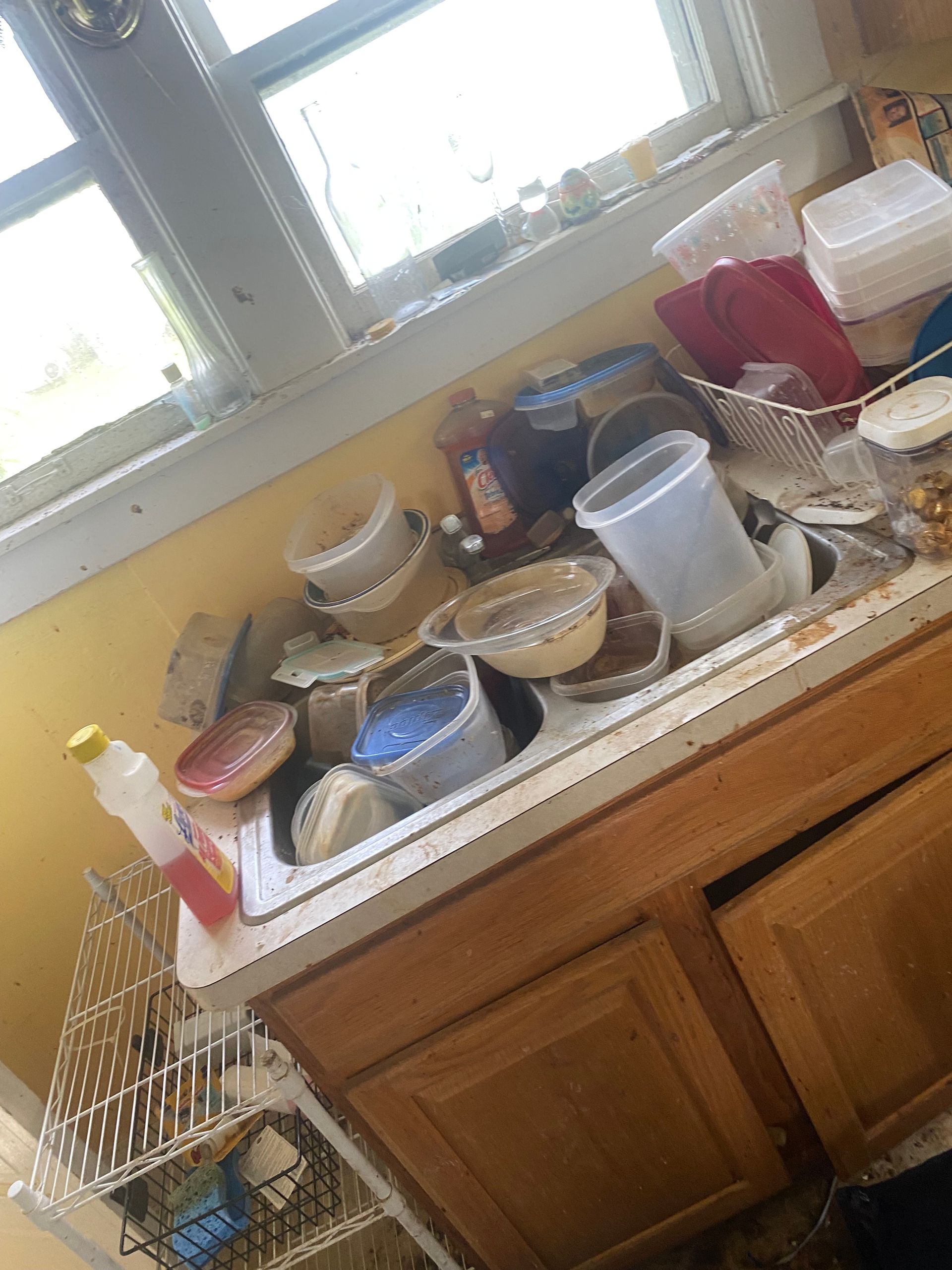 Cluttered kitchen sink with dirty dishes, cups, and containers under a window with sunlight.