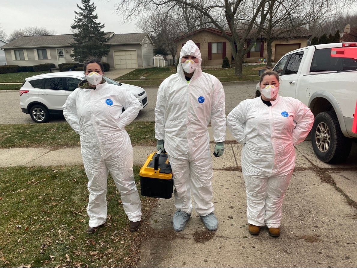 Three people in white protective suits and masks stand on a residential street beside a parked SUV.