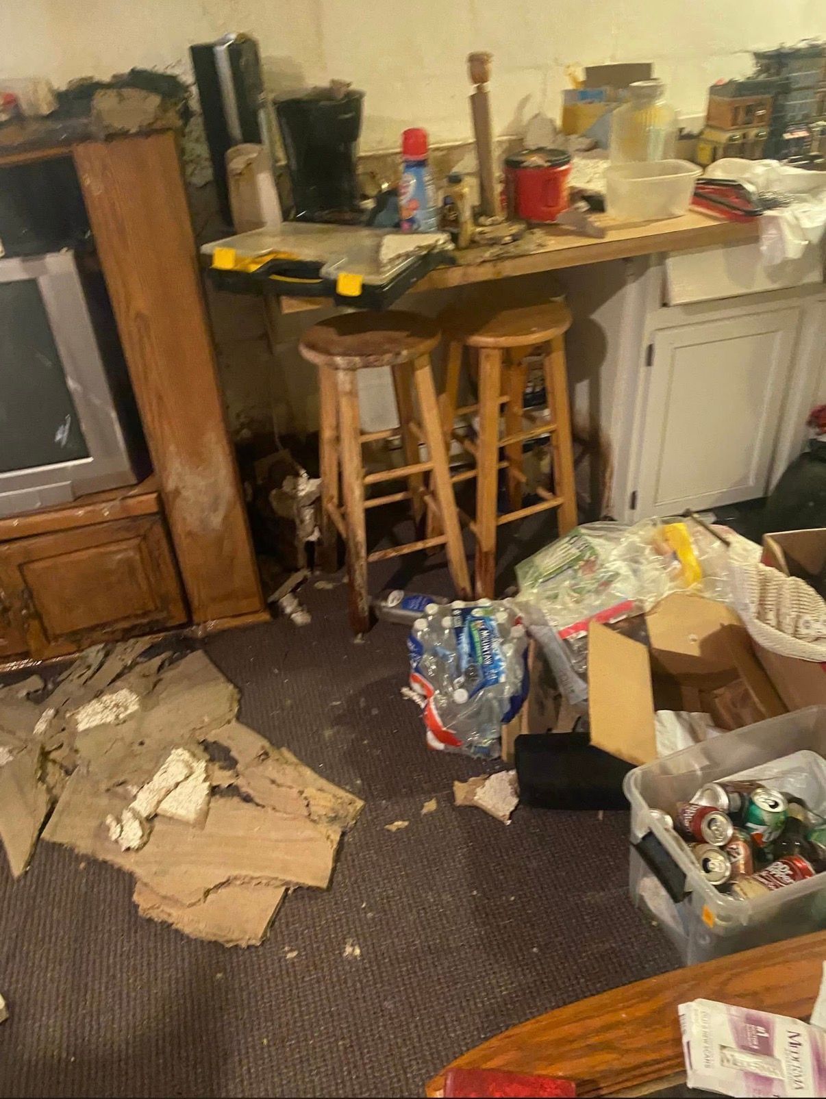 Cluttered kitchen corner with a table, stools, boxes, and items scattered on the floor and counter.