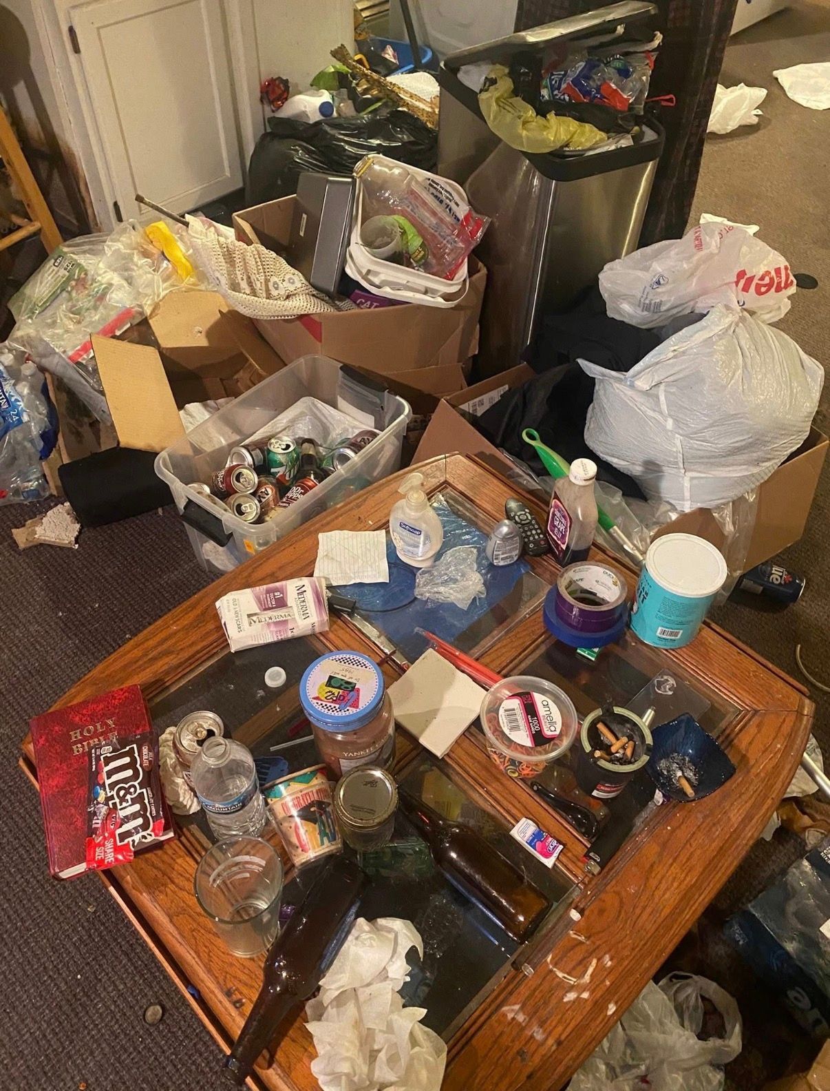 Cluttered room with a messy coffee table covered in cans, bottles, and trash bags in the background