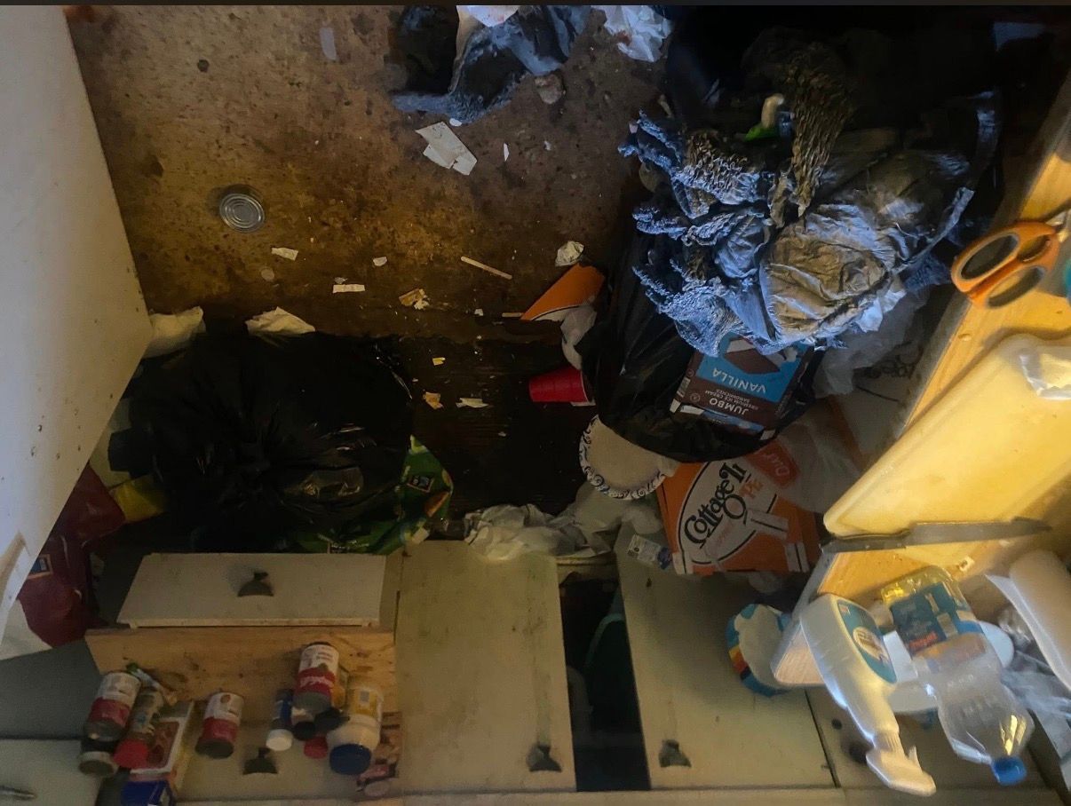 Cluttered floor with cardboard, trash, bags, and scattered bottles in a dim room.