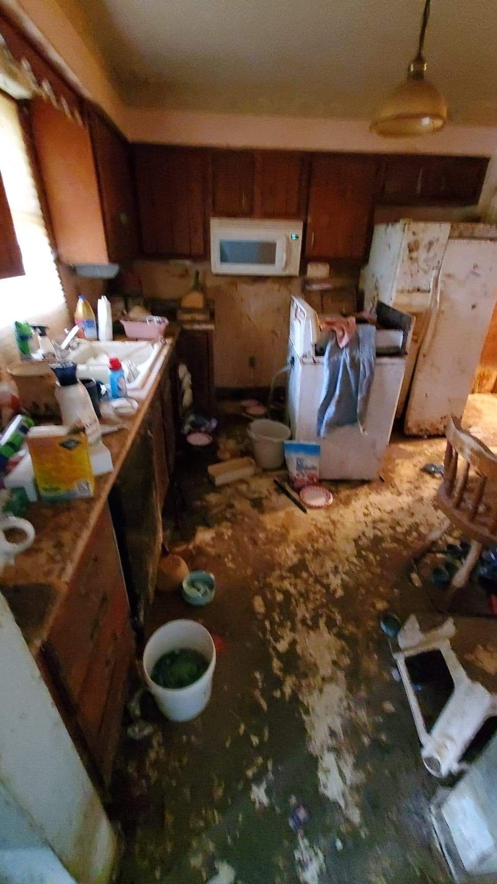Cluttered, dirty kitchen with scattered debris, sink by window, microwave, fridge, and stove.