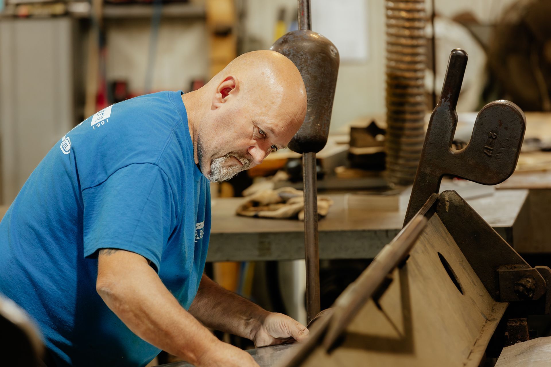 Un homme travaille à la sculpture sur une grande pièce de métal finement ouvragée, dans un atelier. Il est agenouillé et utilise des outils.