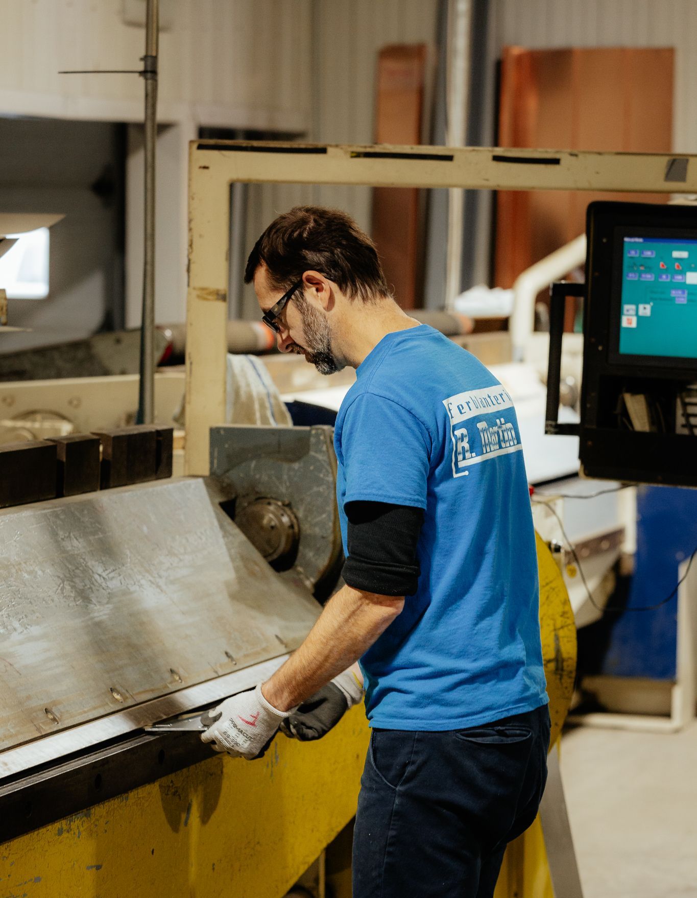 Un homme en chemise bleue travaille le métal dans une usine. Il porte des gants et utilise des machines.