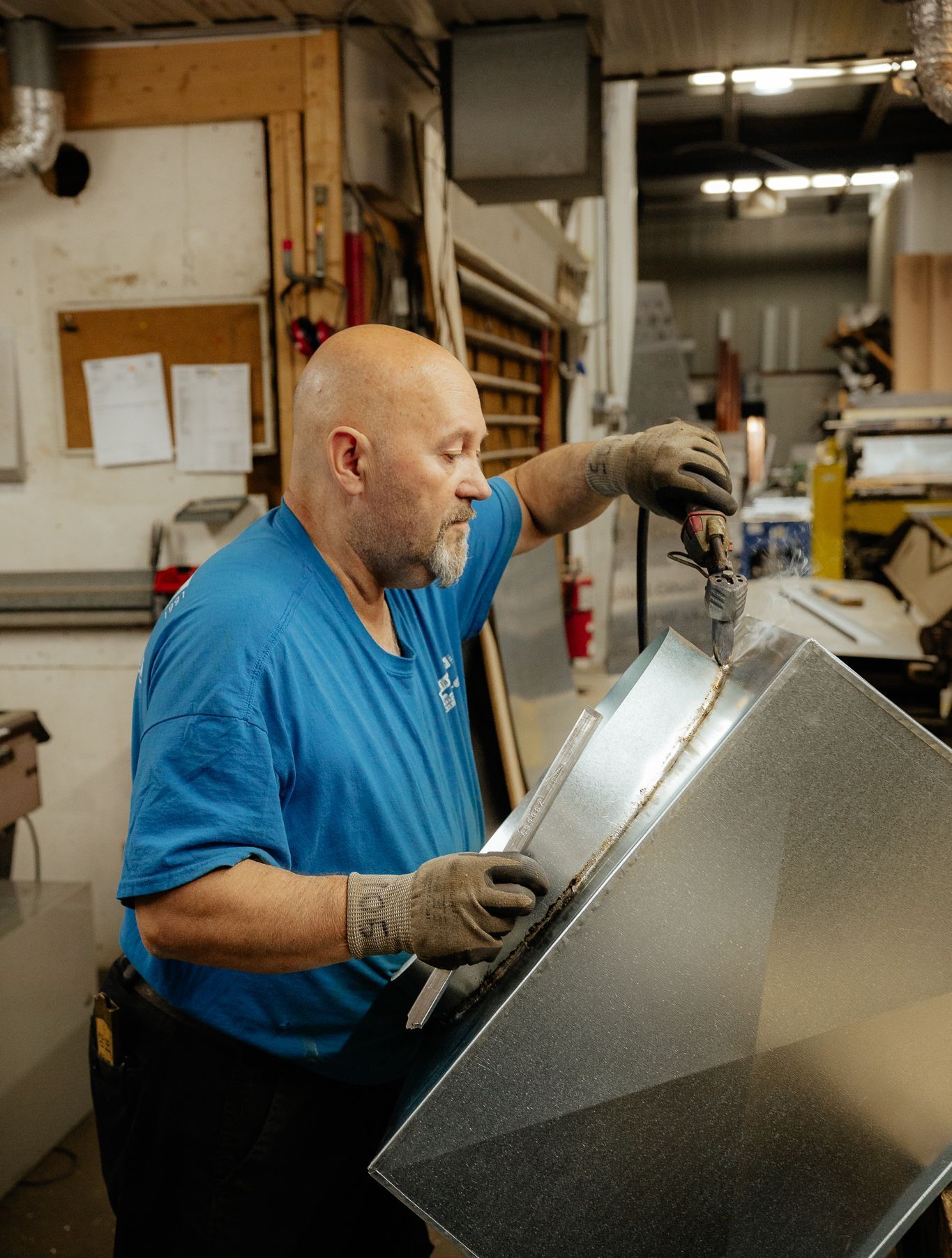 Homme chauve en chemise bleue, travaillant sur des conduits métalliques avec des mains gantées dans un atelier.