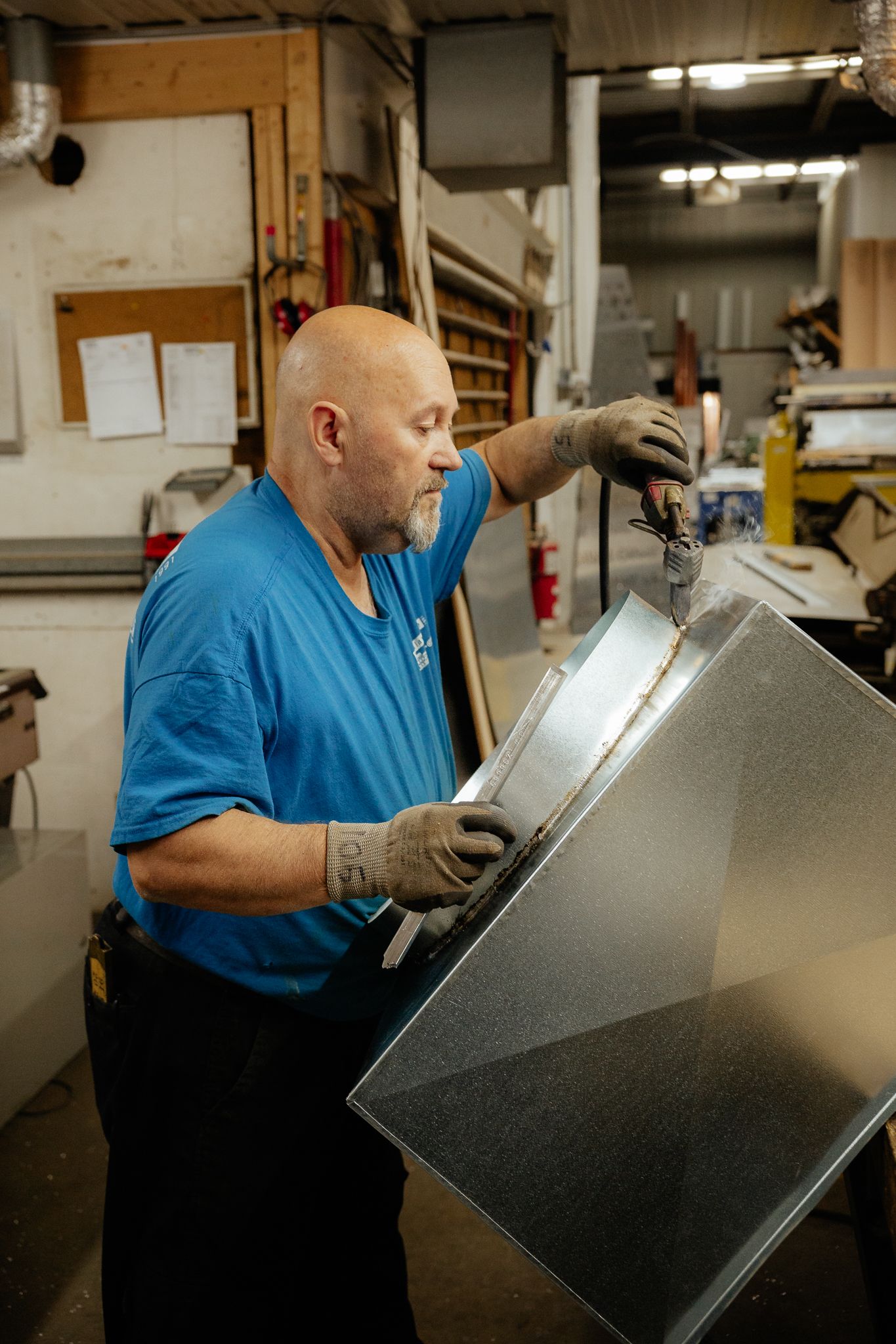 Un homme en chemise bleue travaille la tôle dans un atelier. Il utilise des outils pour l'assembler.