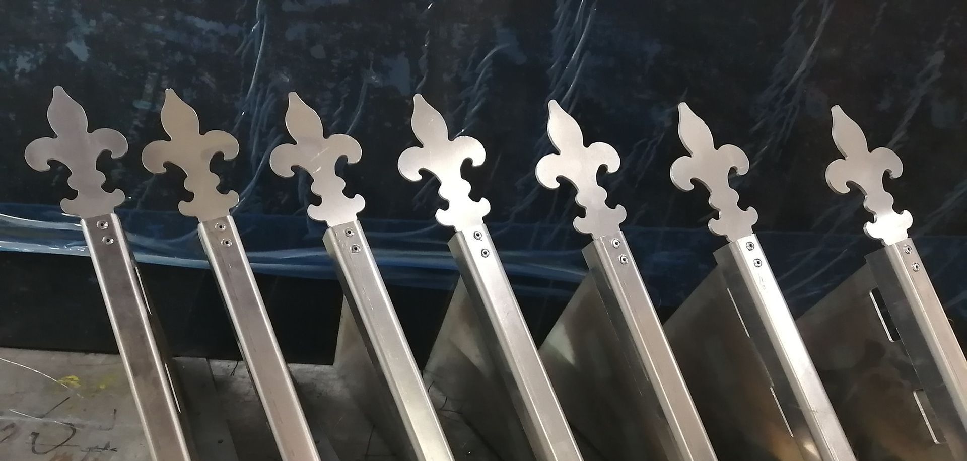 Sept poteaux de clôture en métal à fleurs de lys alignés, légèrement inclinés, à l'intérieur sur un fond sombre.