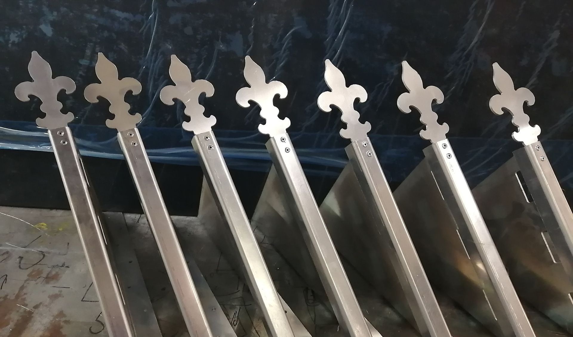 Sept poteaux de clôture en métal avec des sommets en forme de fleur de lys, reposant contre une surface métallique.