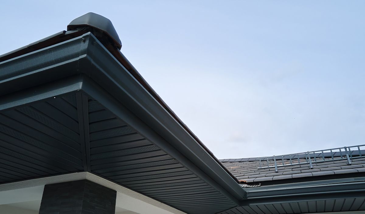 Coin de toit gris foncé avec gouttière et soffite, contre un ciel bleu.