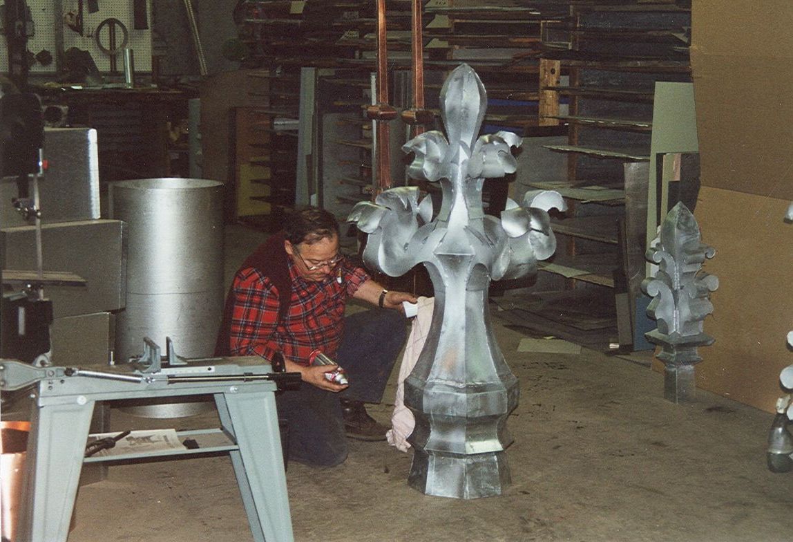 Un homme polit une grande sculpture métallique ornée d'une fleur de lys dans un atelier.