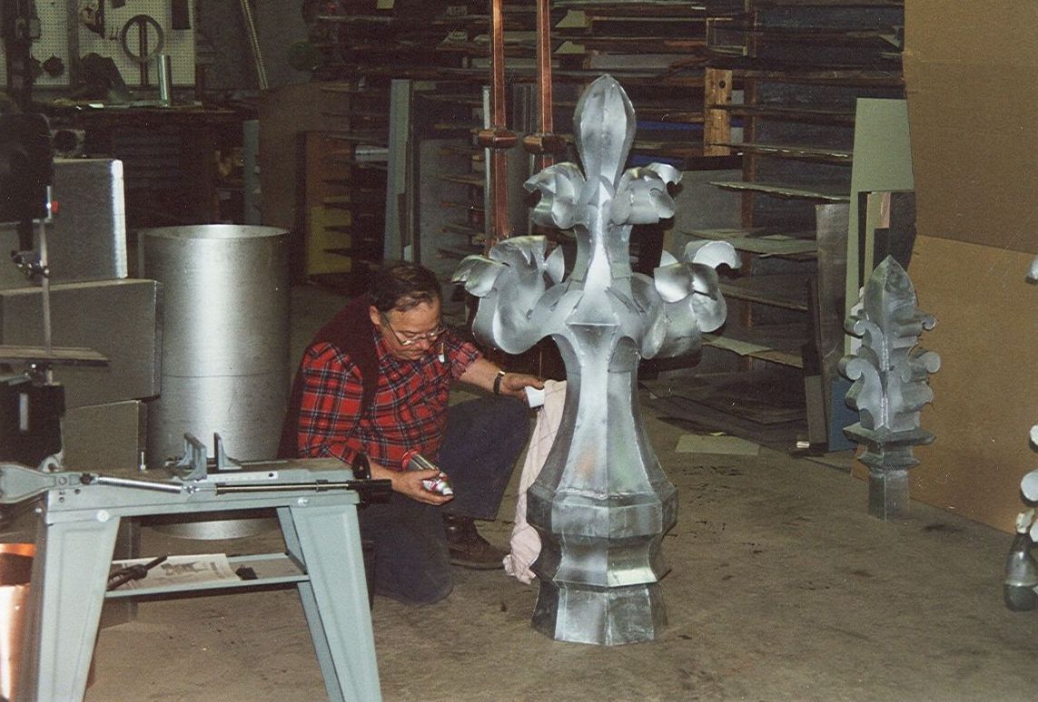 Un homme travaille à la sculpture sur une grande pièce de métal finement ouvragée, dans un atelier. Il est agenouillé et utilise des outils.