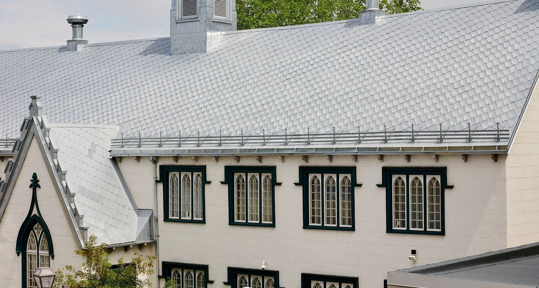 Bâtiment blanc avec toit gris, fenêtres aux garnitures sombres et feuillage vert en arrière-plan.