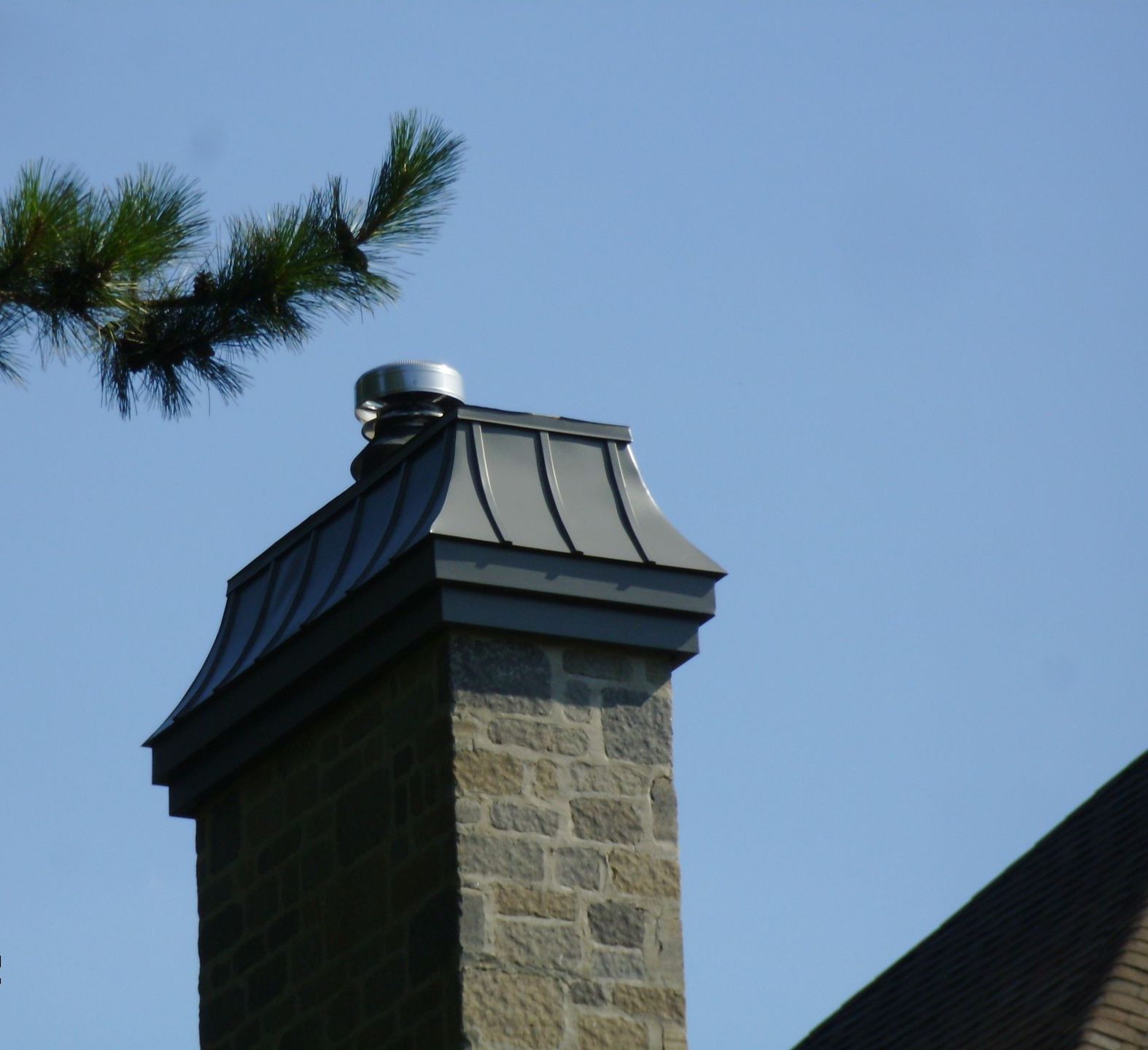 Cheminée en pierre avec chapeau et conduit en métal gris, se détachant sur un ciel bleu.