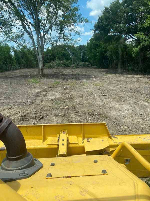 A yellow bulldozer is driving through a dirt field with trees in the background.