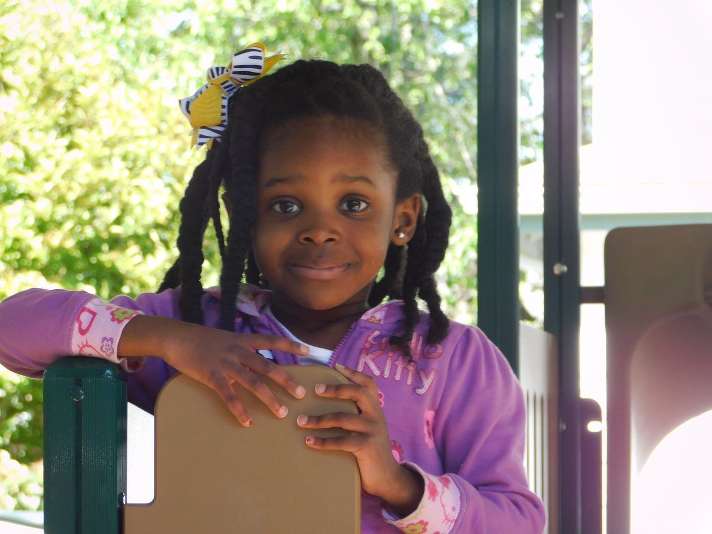 Young girl with locs and bow smiles at camera while leaning on playground structure