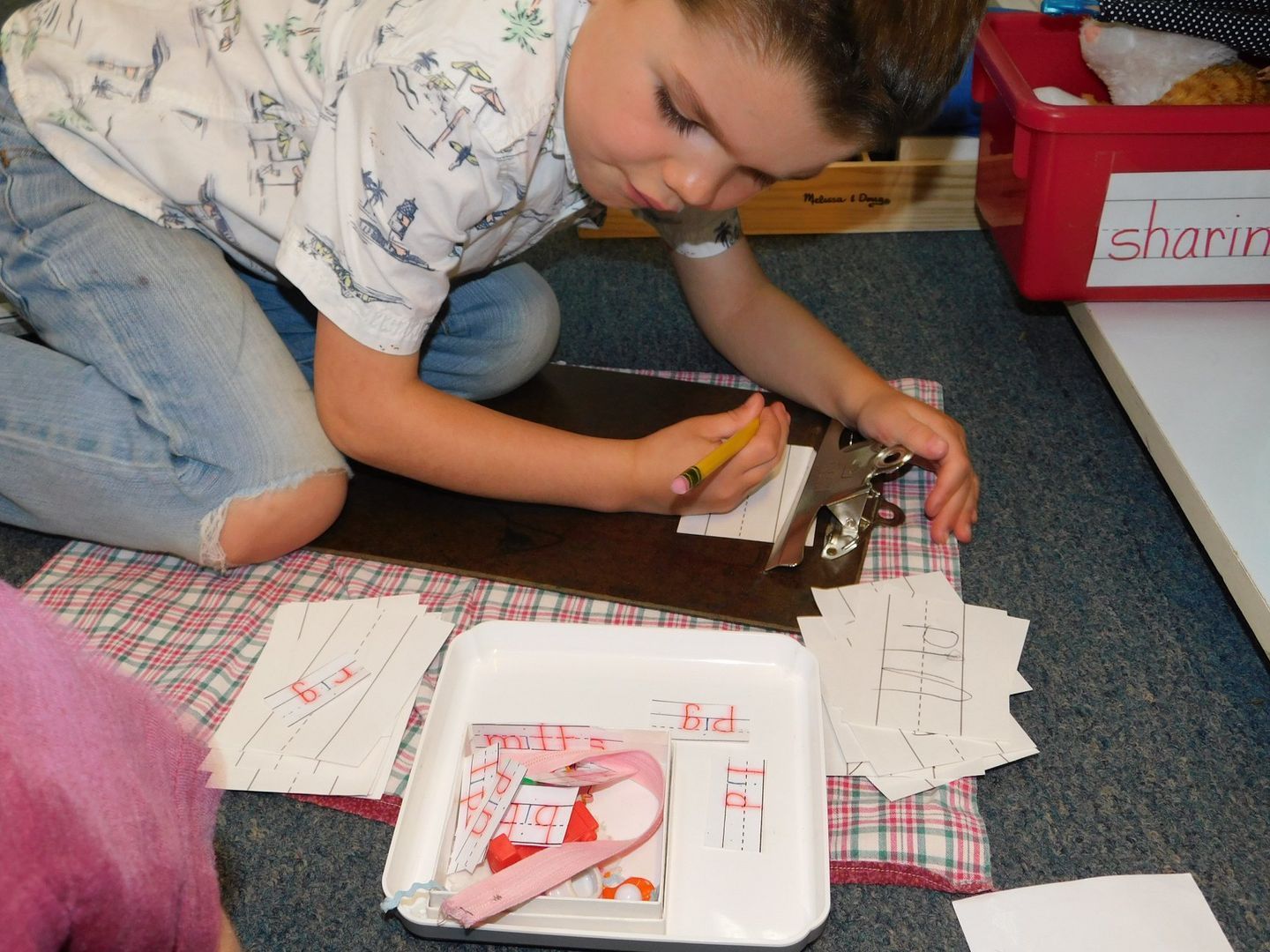 Boy kneeling, writing on clipboard, surrounded by papers and a tray, indoor setting