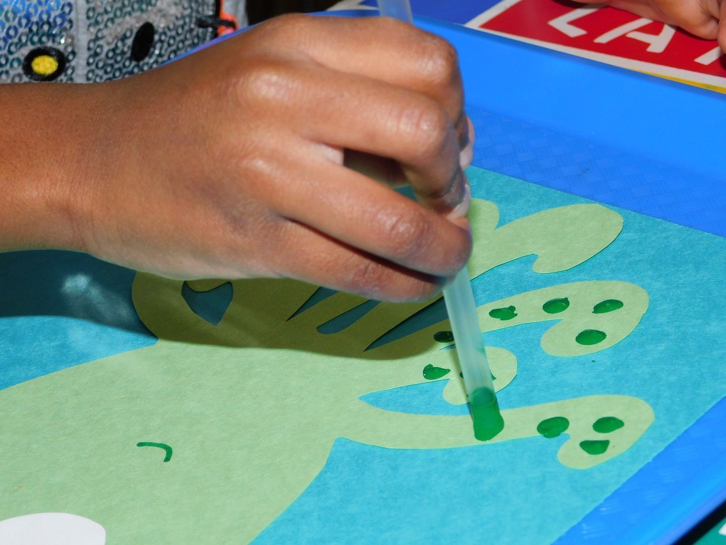 Child's hand using a dropper to apply green paint to a light green paper cutout on a blue tray