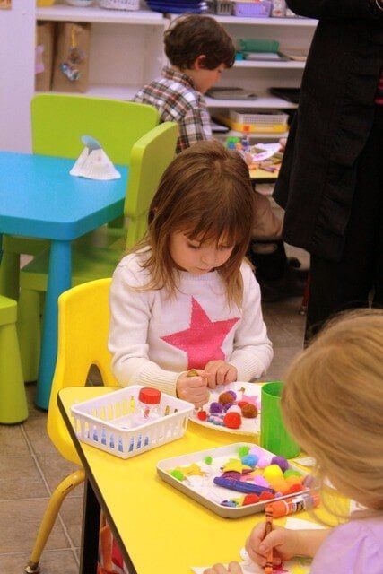 Girl at yellow table, coloring colorful items with other kids in a classroom