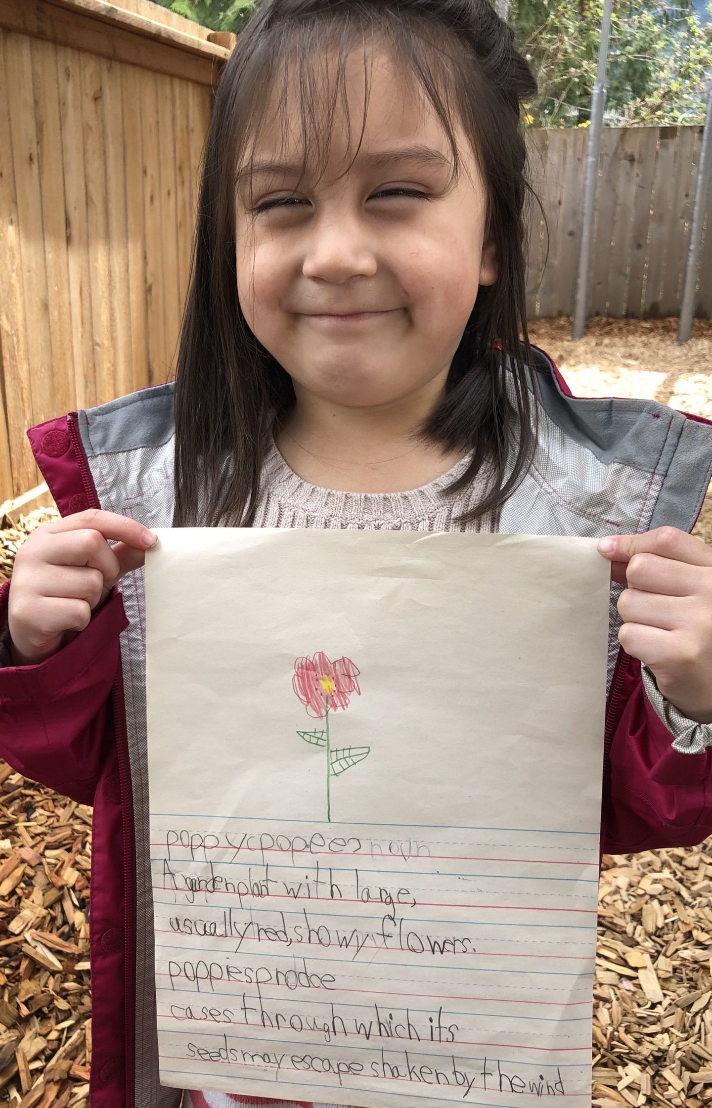 Girl smiles, holding artwork with a drawn flower and text, standing outside near a wooden fence and mulch