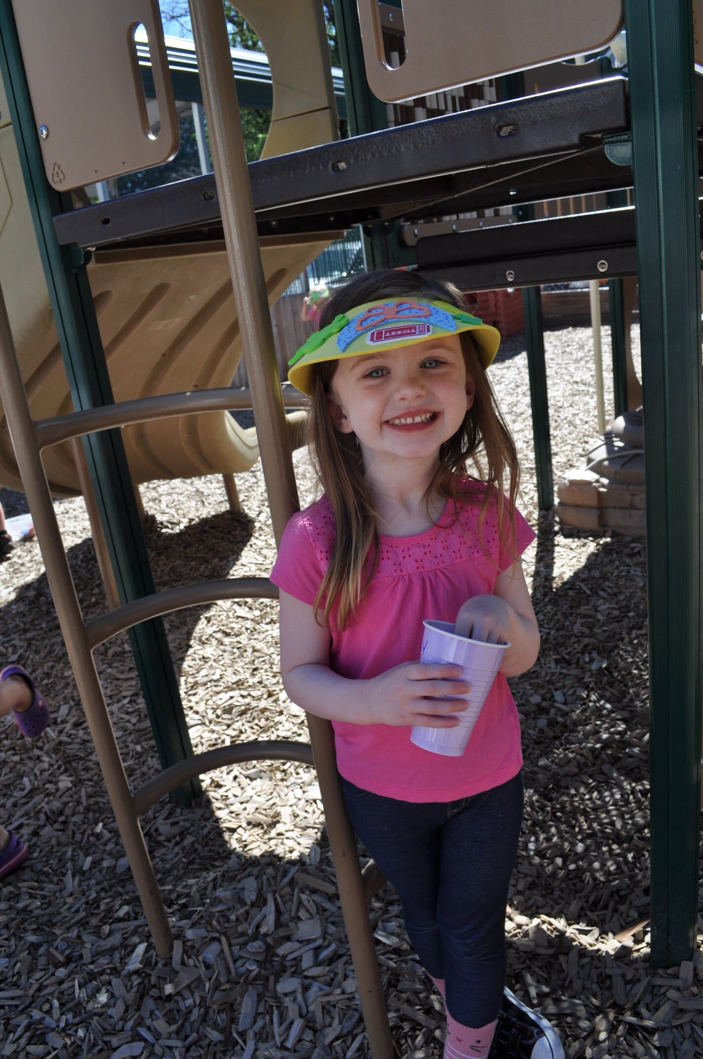 Girl with visor, pink shirt, holding cup, smiling at playground