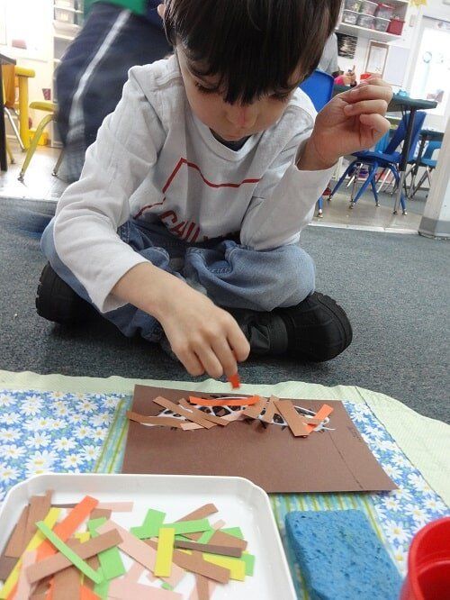 Boy seated, making art with brown, orange, and green paper strips on brown paper