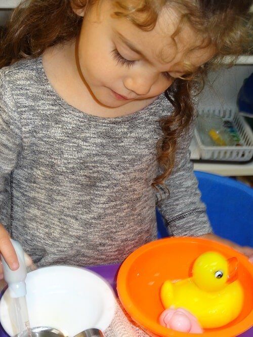 Girl with curly hair playing with a yellow rubber duck, orange bowl, and syringe in a small tub