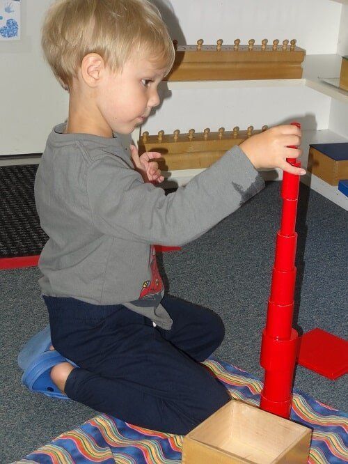 Boy stacking red cylinders, indoors on a patterned mat, with a wooden box