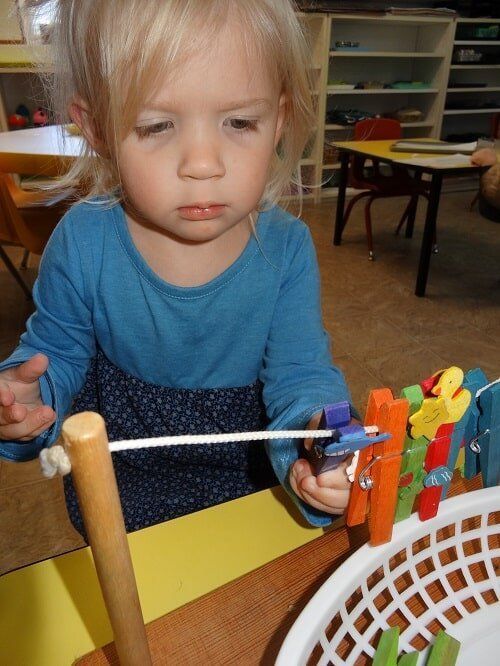 Blond toddler intently working with clothespins, rope, and a wooden stand at a table