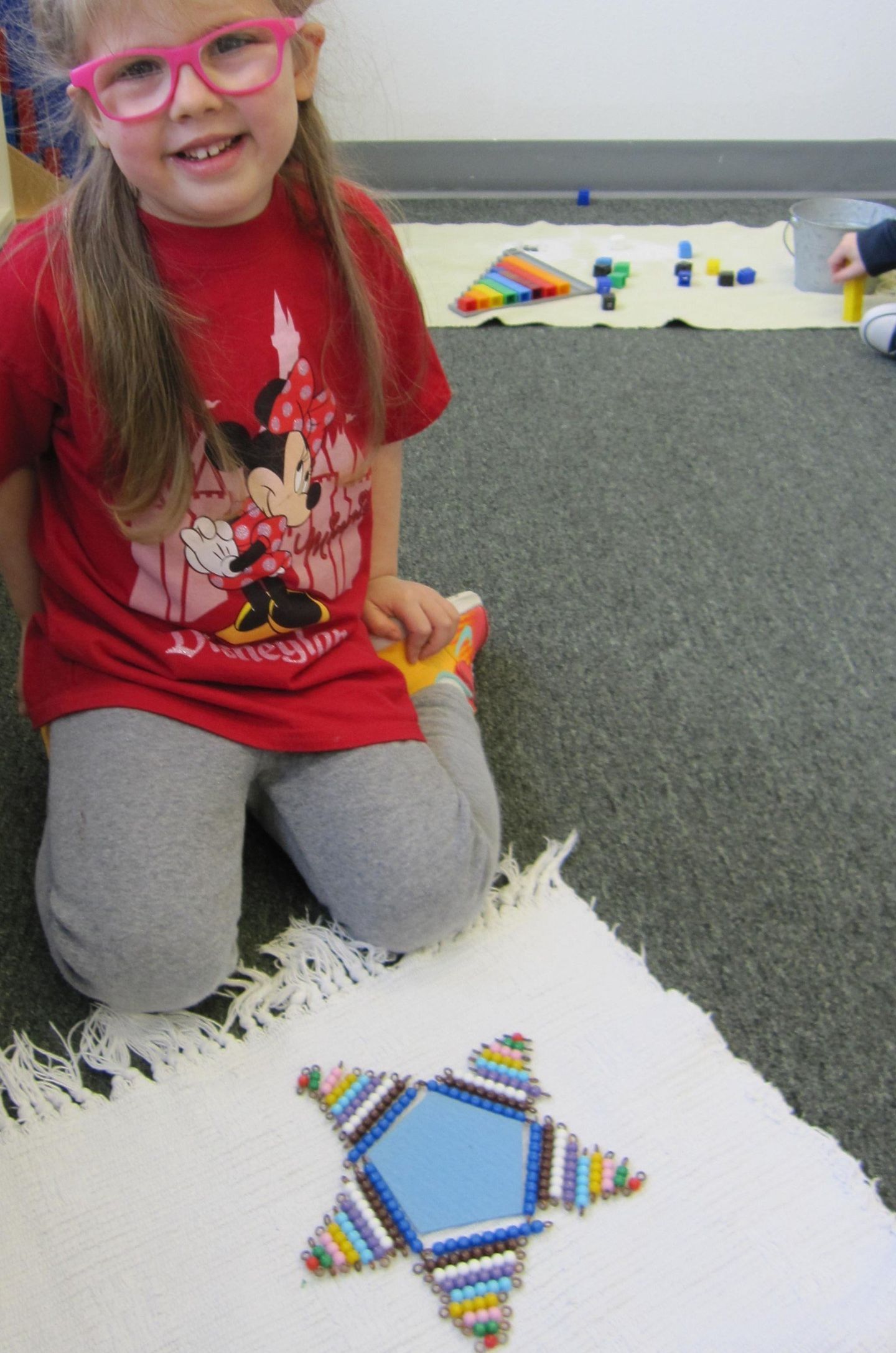 Girl with glasses smiles, kneeling next to a star-shaped craft on a rug