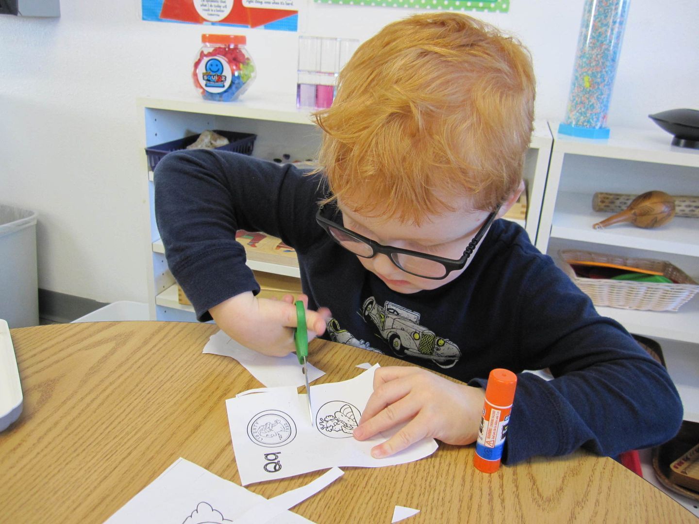 Young child with red hair and glasses cutting paper with scissors at a table