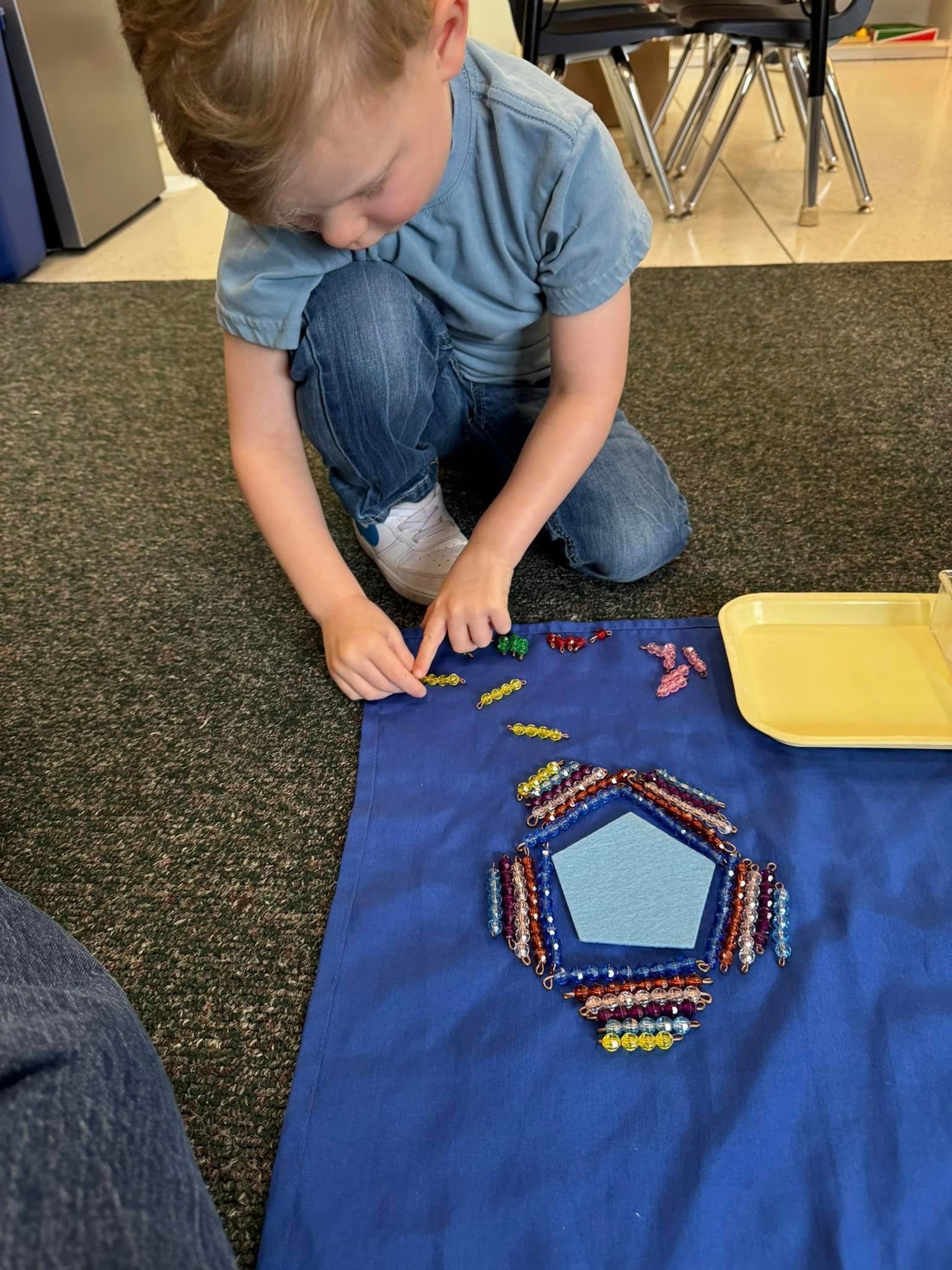 Boy kneeling on carpet arranging colorful beads around a blue pentagon on a blue cloth