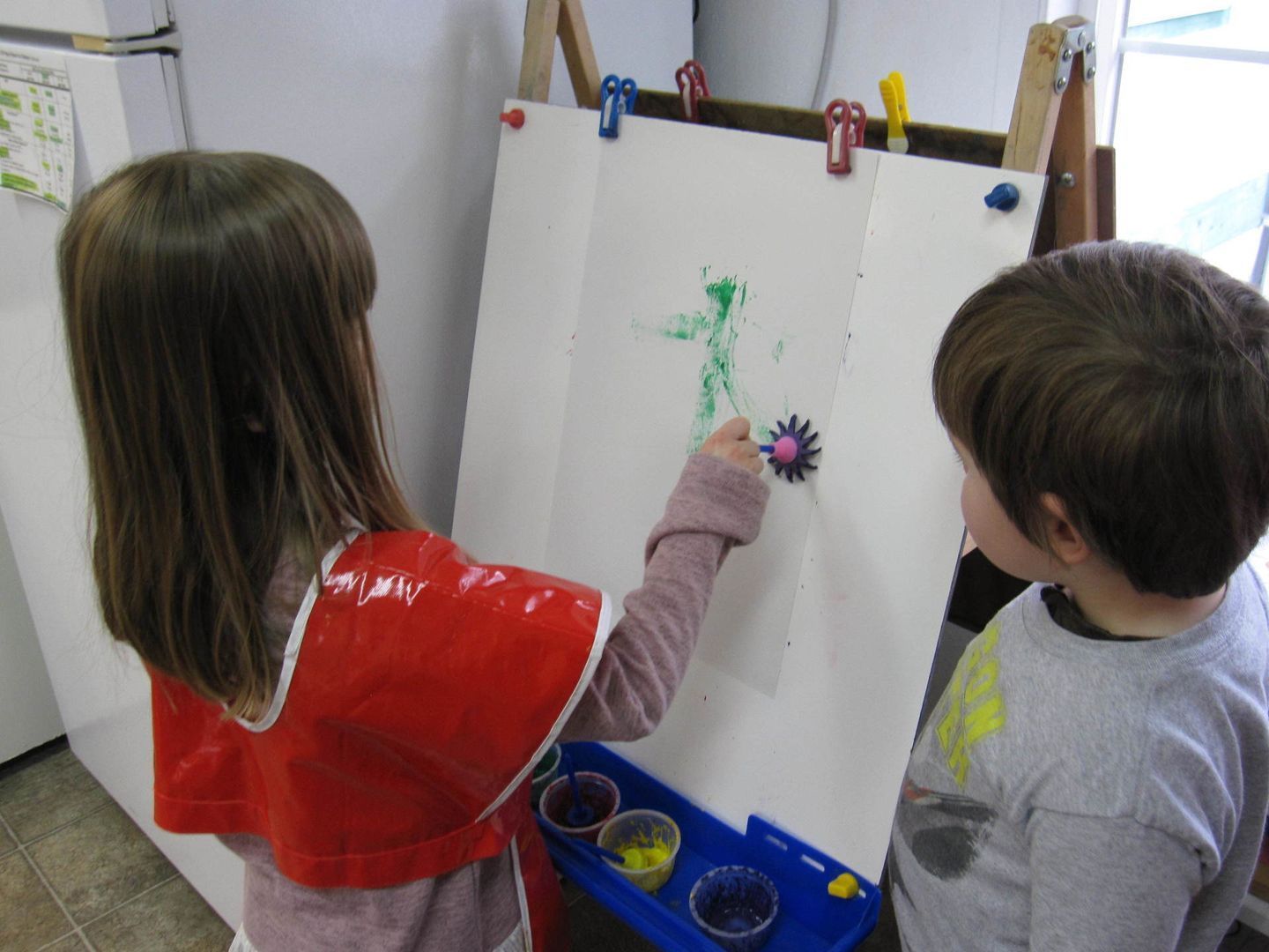 Two children painting at an easel; one paints a flower with green and purple