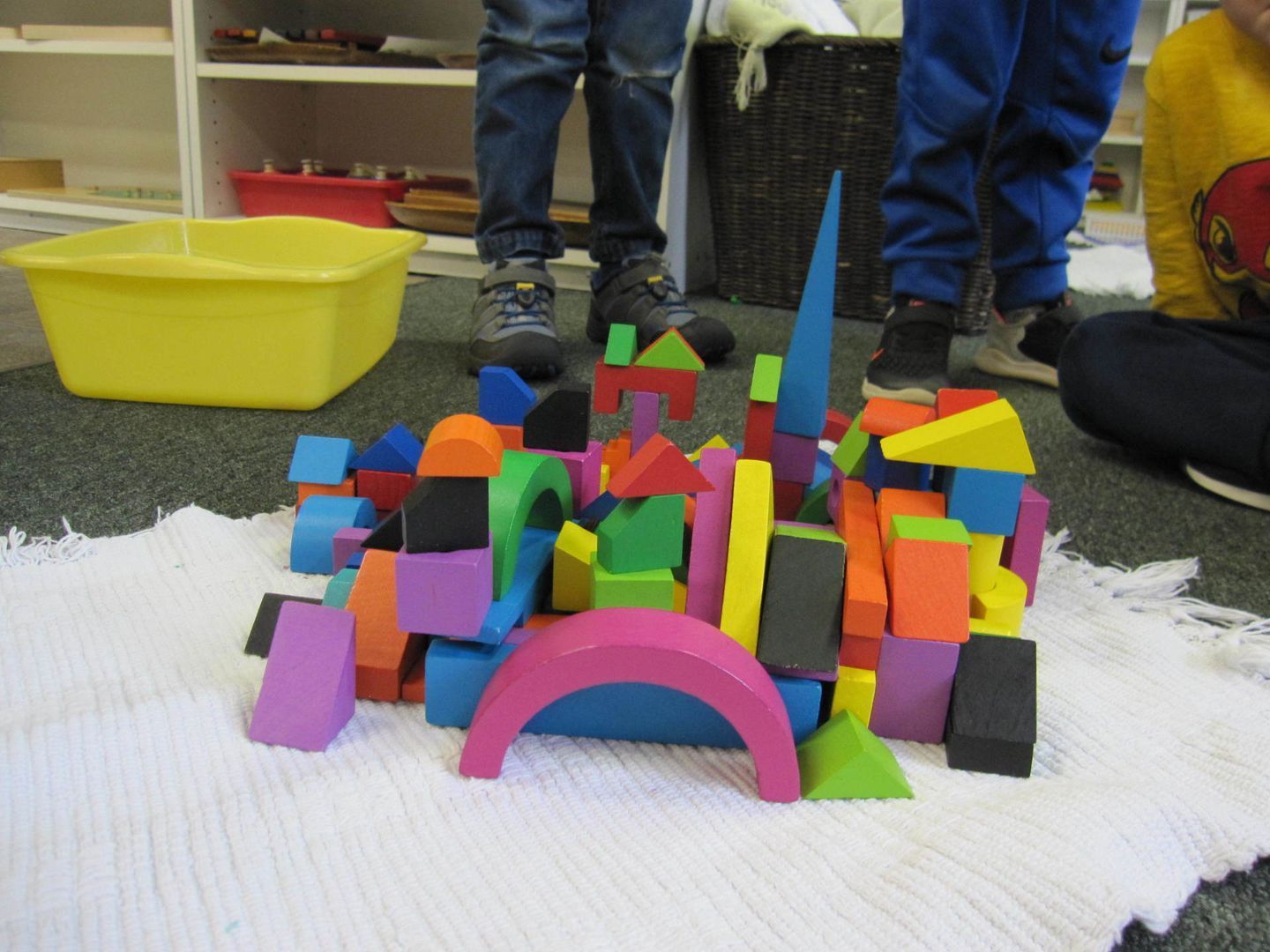 Colorful foam blocks stacked into a structure, children's legs in the background, set on a white rug