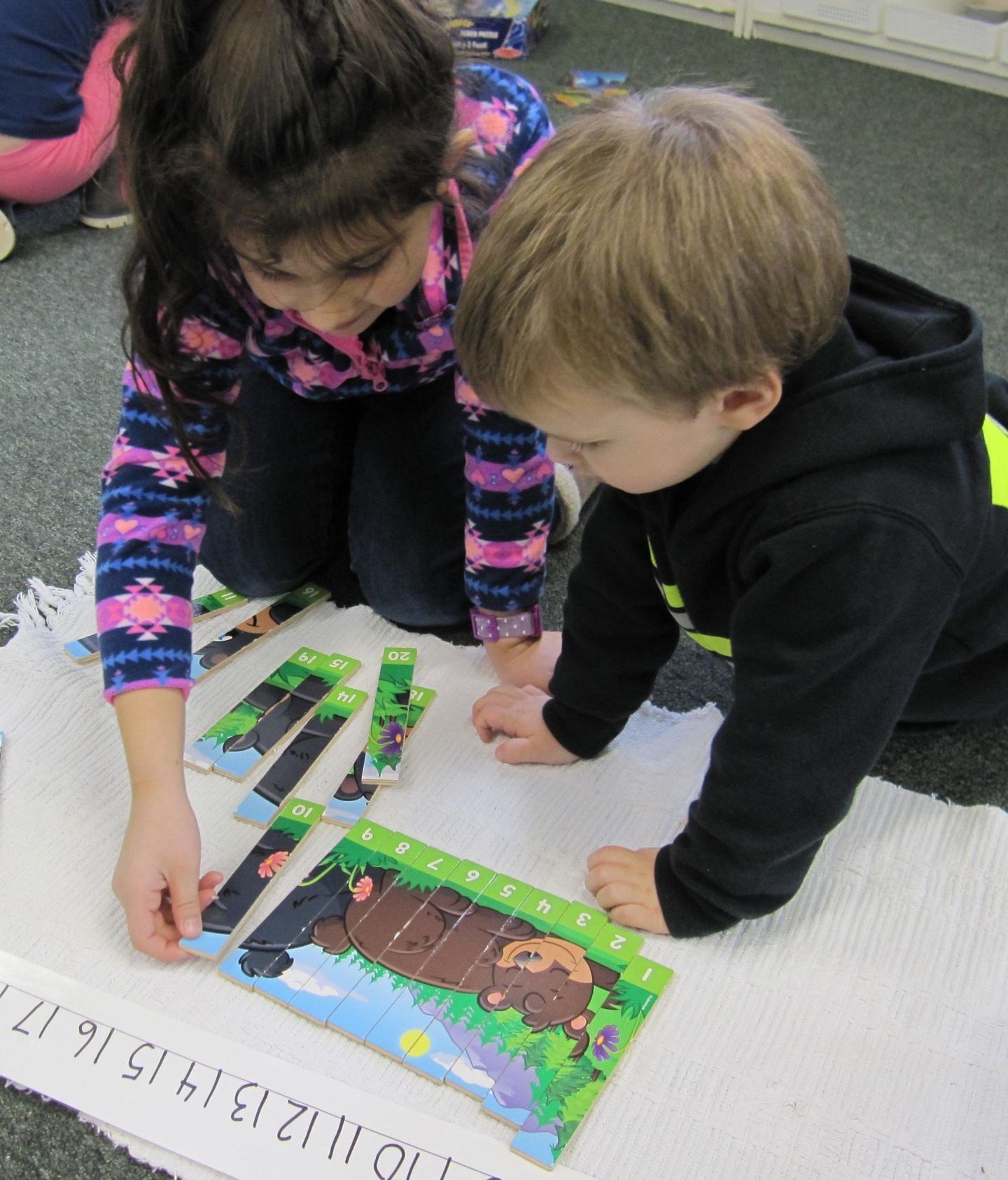 Two children kneel to assemble a puzzle on a white rug