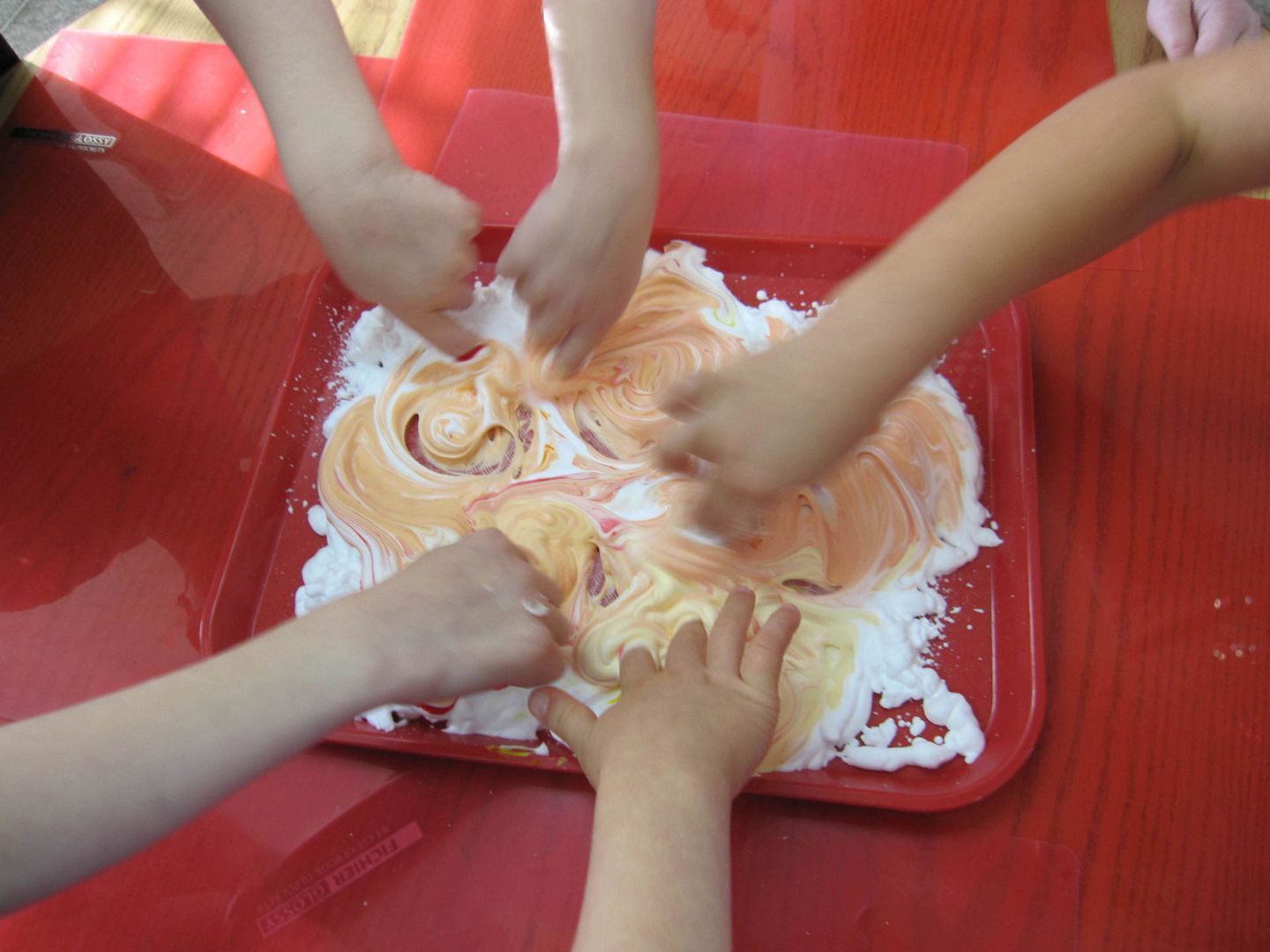 Five children's hands mixing orange and white colors in shaving cream on a red tray