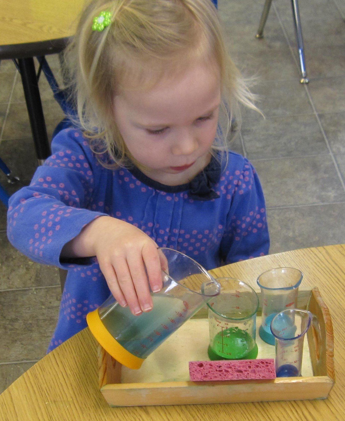 Young girl pouring blue liquid from a pitcher into small glasses on a tray.