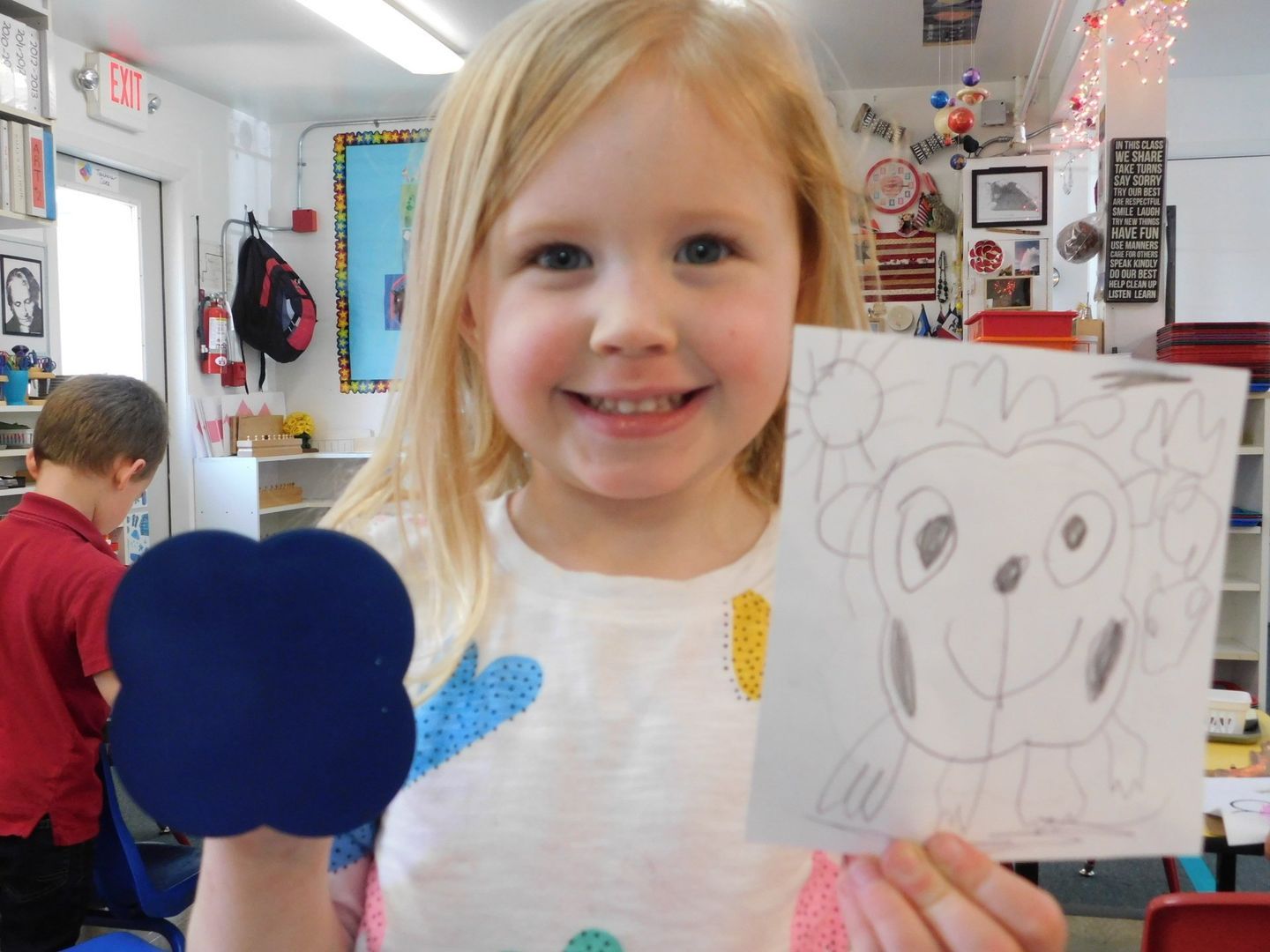 Blonde girl smiles, holding blue shape and monkey drawing in classroom