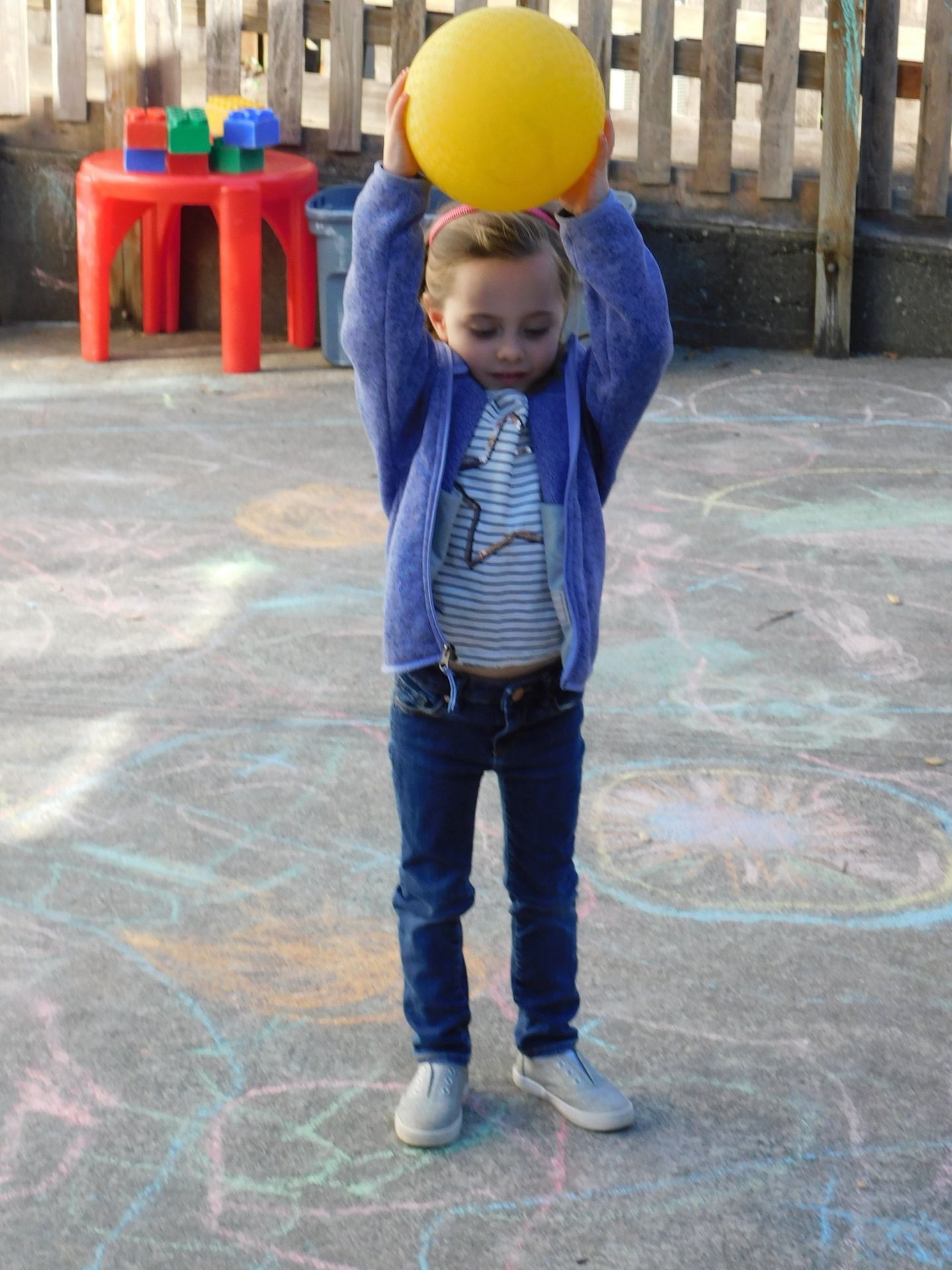 Young girl in blue jacket holding a yellow ball overhead on a chalk-drawn ground